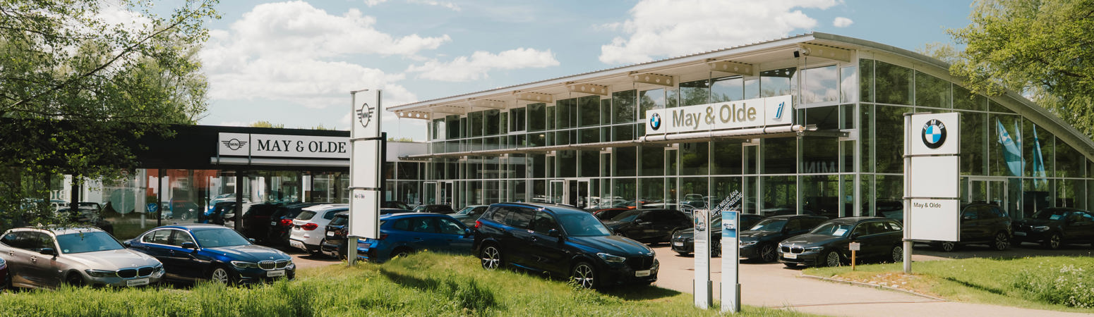 Ein BMW-Autohaus mit mehreren Autos im Freien, einem gläsernen Ausstellungsraum und May & Olde-Schildern unter einem teilweise bewölkten Himmel.