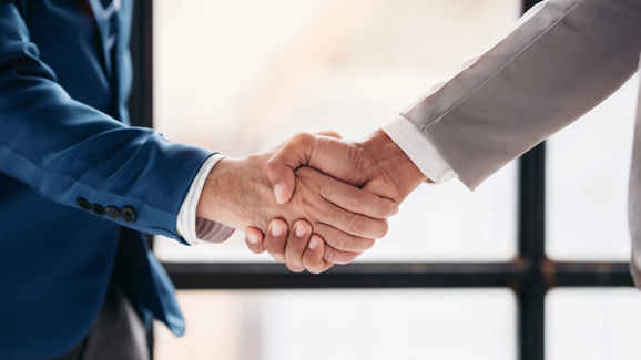 Zwei Männer stehen sich gegenüber. Ihre Hände treffen sich in der Mitte des Bildes, wo diese sich die Hand schütteln.