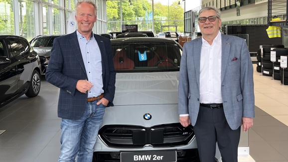 „Zwei Männer stehen in einem Autohaus vor einem grauen BMW 2er mit rotem Innenraum und lächeln in die Kamera.“