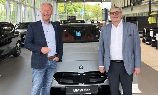 „Zwei Männer stehen in einem Autohaus vor einem grauen BMW 2er mit rotem Innenraum und lächeln in die Kamera.“