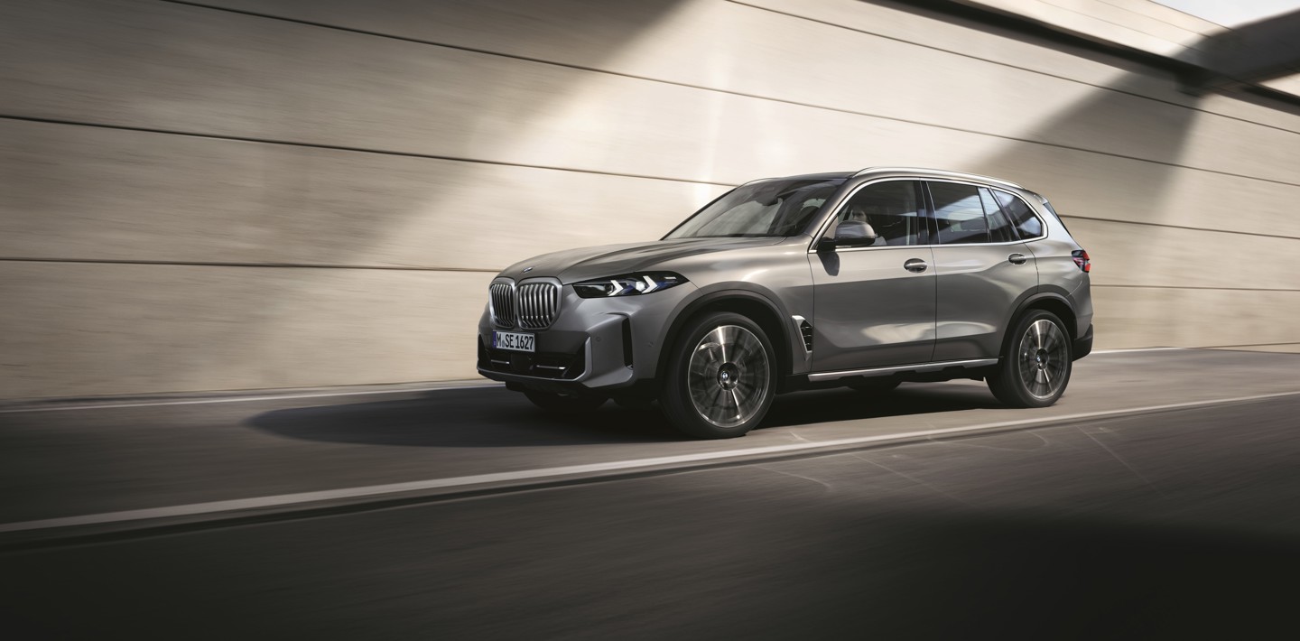 Ein silberner BMW X5 fährt auf einer glatten, sonnenbeschienenen Straße neben einer beigen Betonwand entlang.