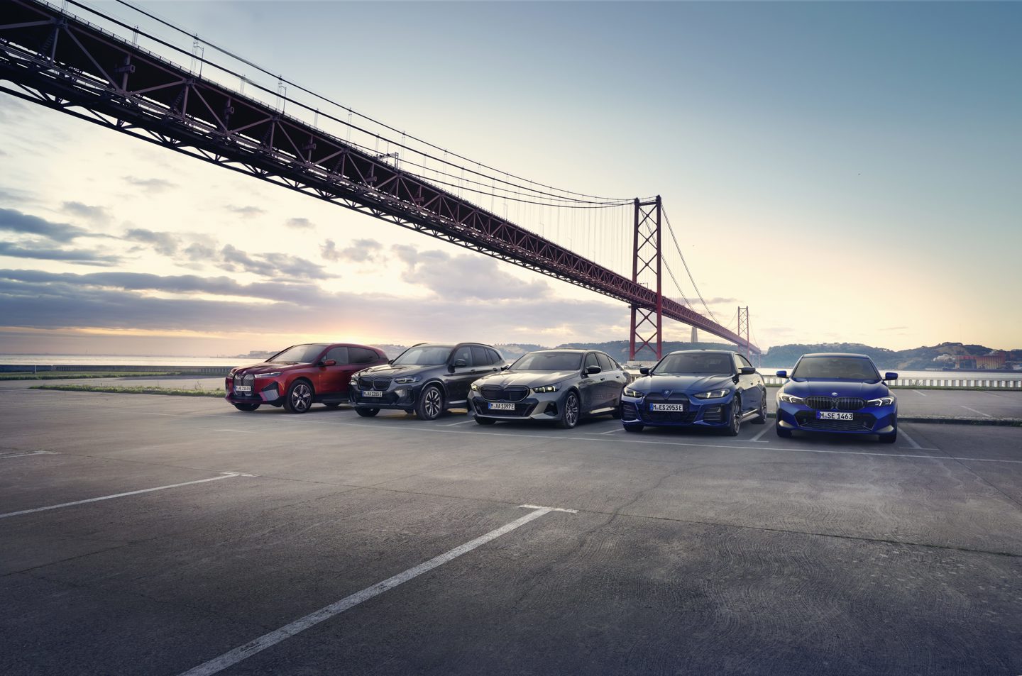 Mehrere BMW Junge Gebrauchtwagen stehen nebeneinander auf einem Parkplatz vor einer Brücke – Kampagnenmotiv der May & Olde Junge Gebrauchtwagen Aktion.