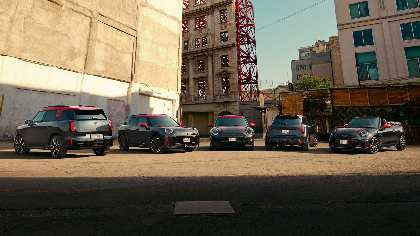 Fünf Mini John Cooper Works mit roten Dächern sind in einer Reihe auf einem städtischen Parkplatz geparkt, im Hintergrund sind Gebäude und Baugerüste zu sehen.
