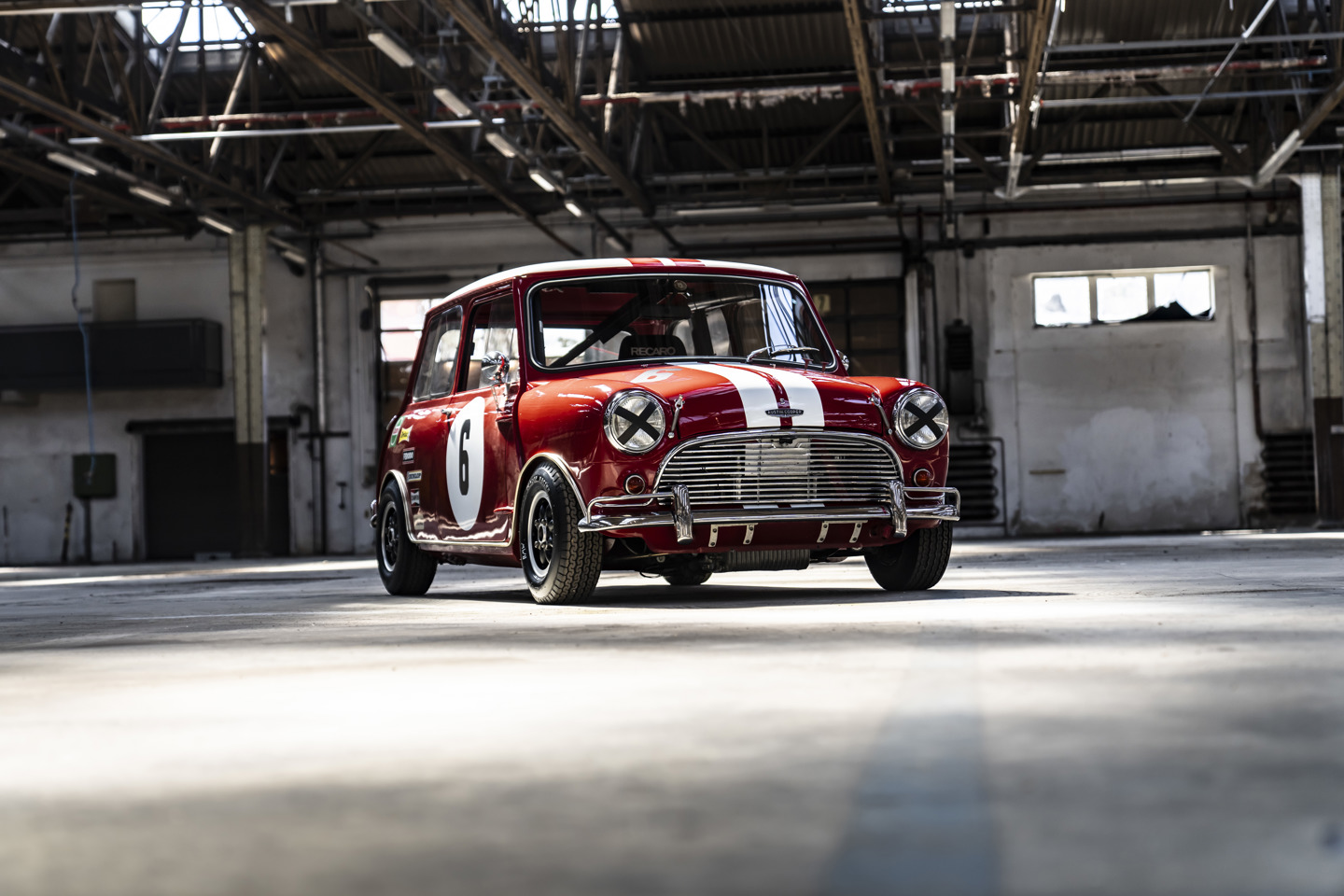 Ein roter Mini Cooper im Vintage-Stil mit Rennstreifen und Startnummernaufklebern steht in einer leeren, sonnenbeschienenen Industriehalle.