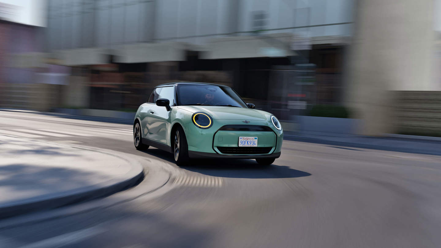 Ein Mintfarbender vollelektrischer MINI Cooper E fährt um eine Straßenecke in der Stadt, wobei der Hintergrund ein Gefühl der Bewegung vermittelt.