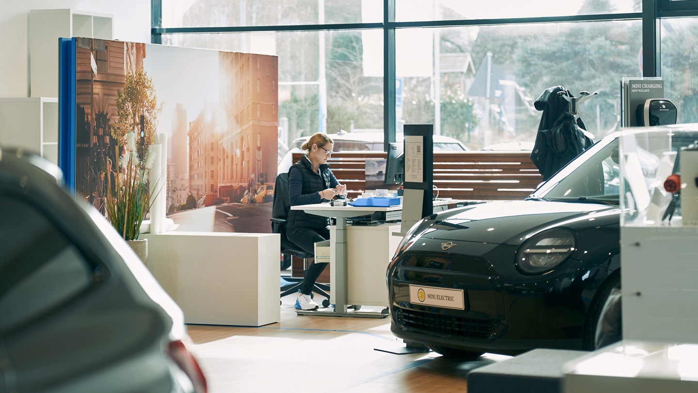 „Innenaufnahme eines modernen MINI Autohauses. Im Vordergrund steht ein schwarzer MINI Electric, daneben eine Service-Mitarbeiterin am Schreibtisch in Beratungssituation. Im Hintergrund ein großes Wandbild mit urbanem Straßenzug, moderne Einrichtung und klare Tageslicht-Atmosphäre.“