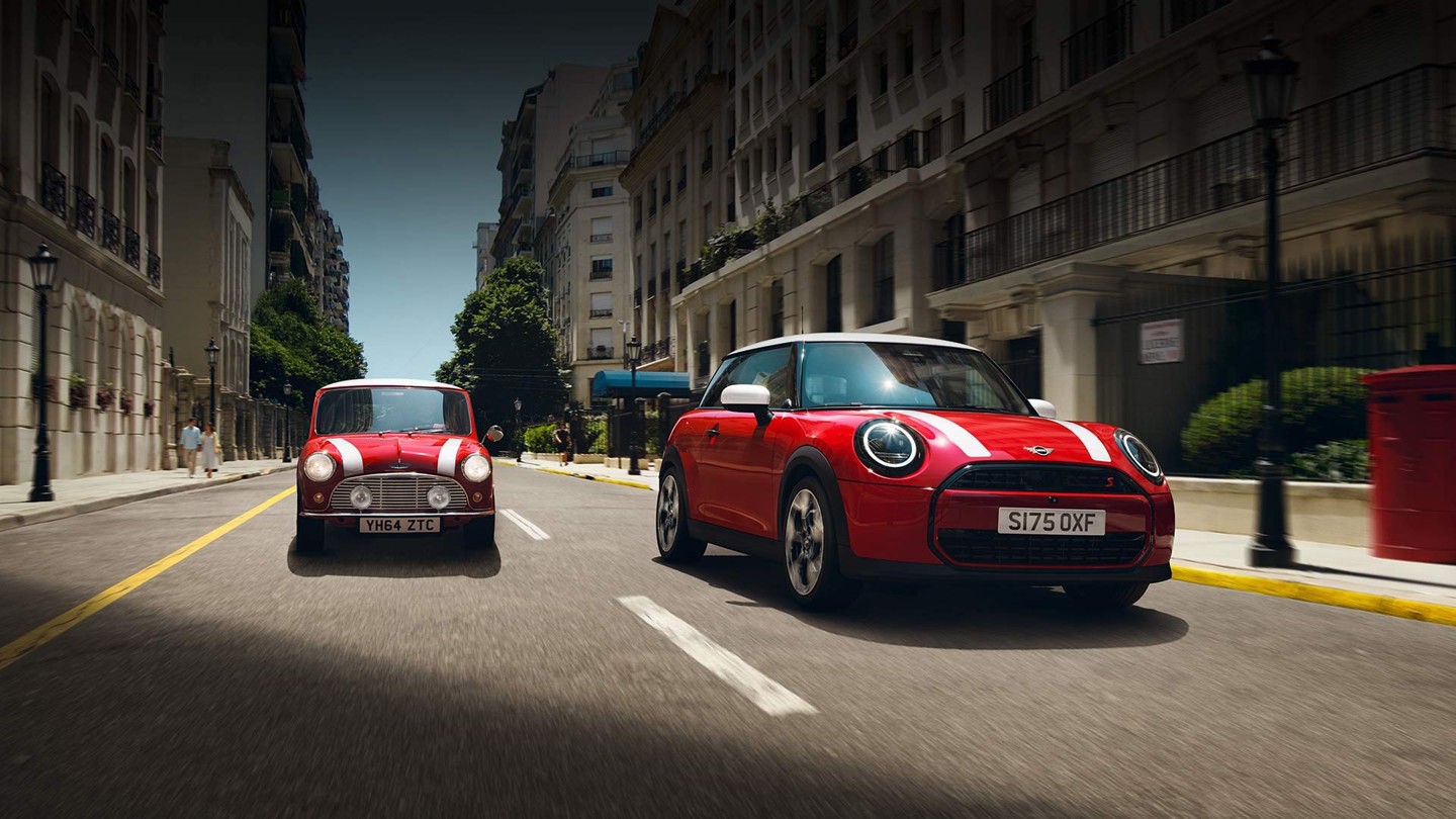 Zwei rote MINI Fahrzeuge, ein klassischer MINI Oldtimer und ein moderner MINI Cooper, fahren nebeneinander auf einer städtischen Straße mit historischer Architektur im Hintergrund, dynamische Fahrszene mit Fokus auf Designentwicklung, Tradition und modernes Fahrgefühl ohne eingeblendeten Werbetext