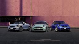 rei MINI Modelle – ein silbernes Cabrio, ein mintgrüner Dreitürer und ein blauer Fünftürer – nebeneinander geparkt vor einer modernen pinkfarbenen Gebäudefassade bei Tageslicht.