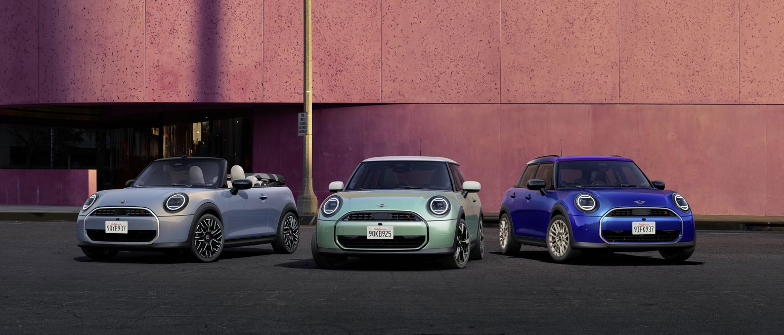 rei MINI Modelle – ein silbernes Cabrio, ein mintgrüner Dreitürer und ein blauer Fünftürer – nebeneinander geparkt vor einer modernen pinkfarbenen Gebäudefassade bei Tageslicht.