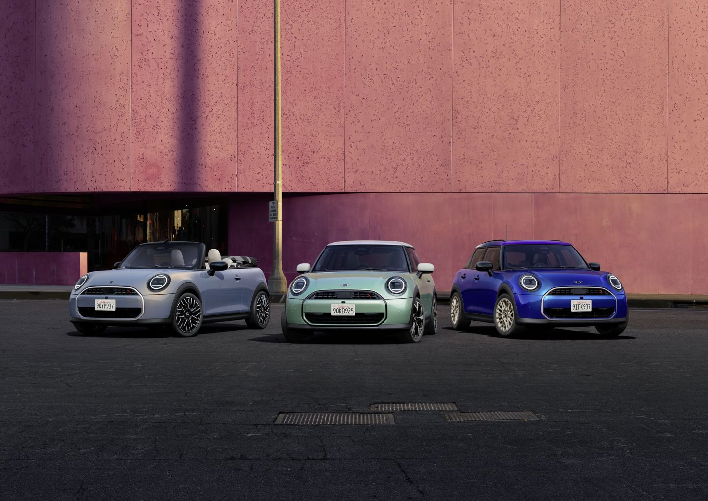 rei MINI Modelle – ein silbernes Cabrio, ein mintgrüner Dreitürer und ein blauer Fünftürer – nebeneinander geparkt vor einer modernen pinkfarbenen Gebäudefassade bei Tageslicht.