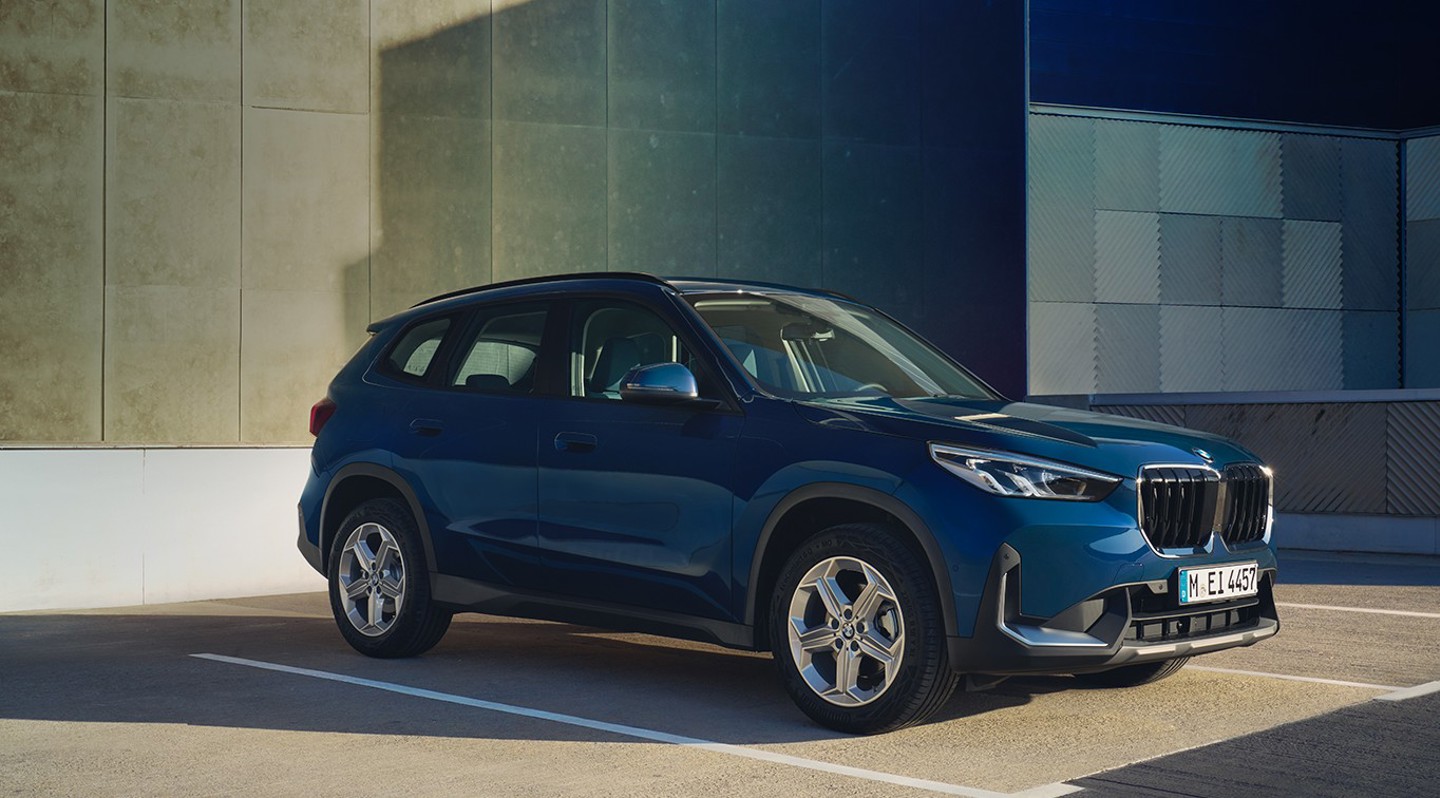 Ein blauer BMW X1 steht auf einem Parkplatz im Freien neben einem modernen Gebäude mit gemusterten Wänden.