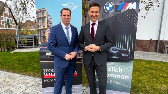 „Zwei Männer in Anzügen stehen nebeneinander vor zwei Roll-Up-Bannern mit BMW und MINI Logos. Im Hintergrund Fachwerkhaus und moderner Gebäudeteil.“