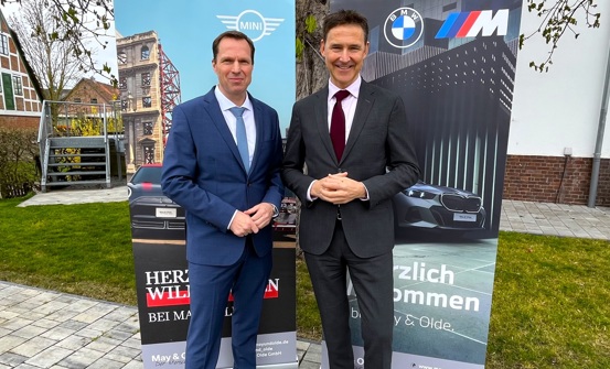 „Zwei Männer in Anzügen stehen nebeneinander vor zwei Roll-Up-Bannern mit BMW und MINI Logos. Im Hintergrund Fachwerkhaus und moderner Gebäudeteil.“