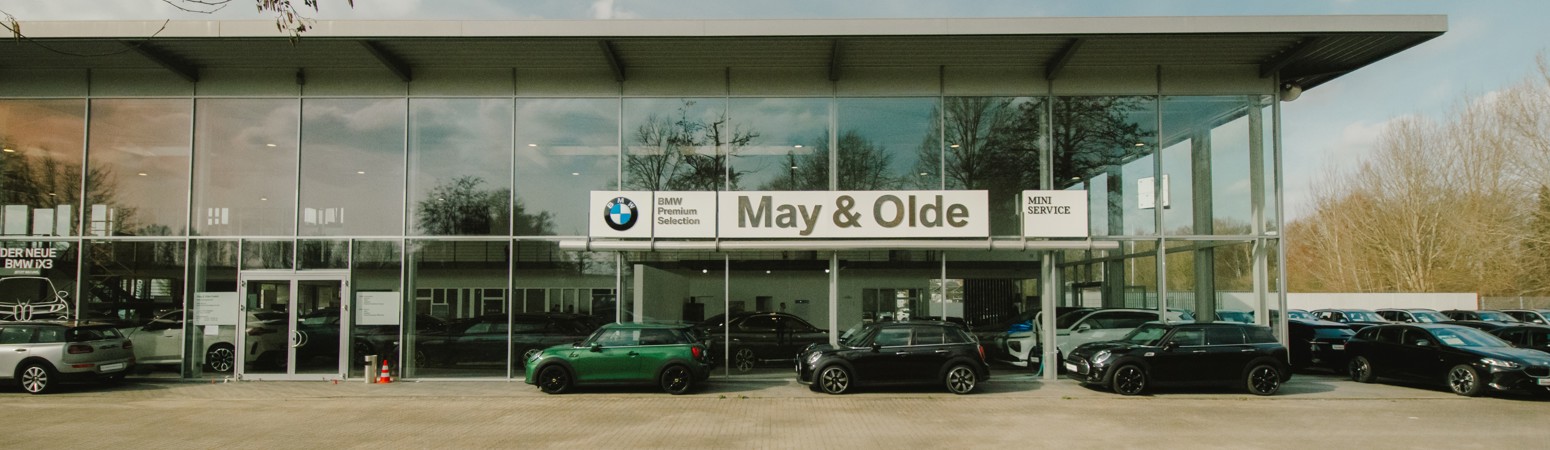 Ein BMW-Händler mit einem May & Olde-Schild, auf dem mehrere Autos vor einem glatten Glasgebäude an einem hellen, klaren Tag abgebildet sind.