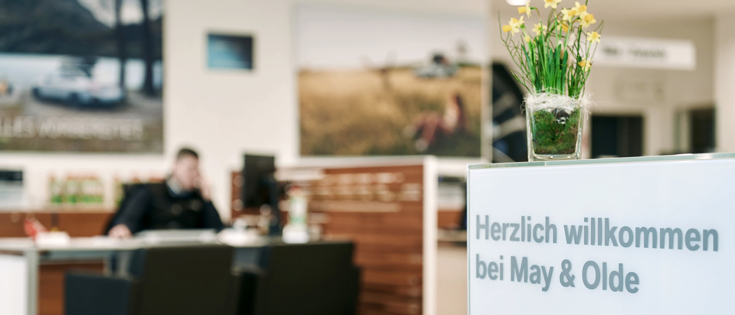 „Innenaufnahme eines Autohauses von May & Olde mit Fokus auf ein Begrüßungsschild mit der Aufschrift ‚Herzlich willkommen bei May & Olde‘ und einem Blumentopf mit gelben Narzissen. Im Hintergrund ein unscharfer Mitarbeiter an einem Schreibtisch.“