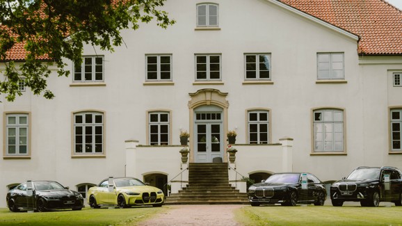 Vier BMW Luxusautos, darunter ein BMW M4 Competition Cabrio, ein BMW M4 CS, ein BMW i7 und ein BMW X7 stehen auf dem Rasen vor einem großen, weißen, zweistöckigen Gebäude mit rotem Ziegeldach und vielen Fenstern.