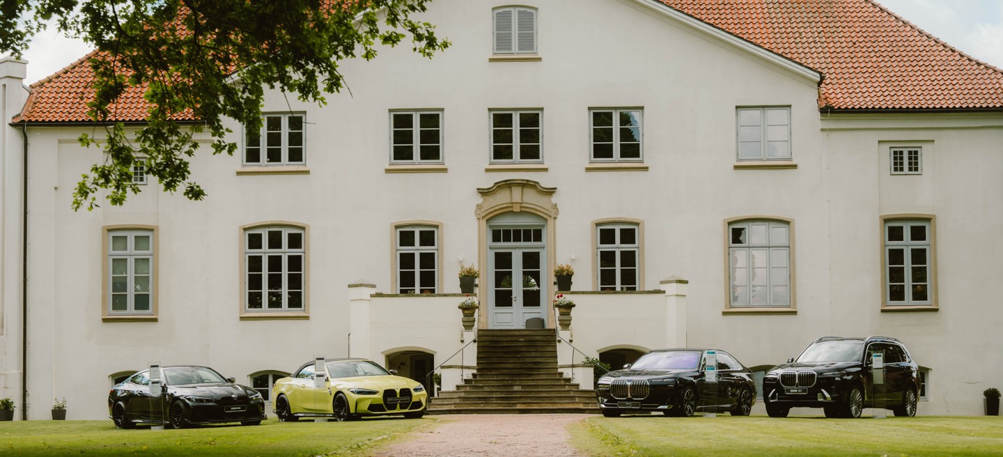 Vier BMW Luxusautos, darunter ein BMW M4 Competition Cabrio, ein BMW M4 CS, ein BMW i7 und ein BMW X7 stehen auf dem Rasen vor einem großen, weißen, zweistöckigen Gebäude mit rotem Ziegeldach und vielen Fenstern.