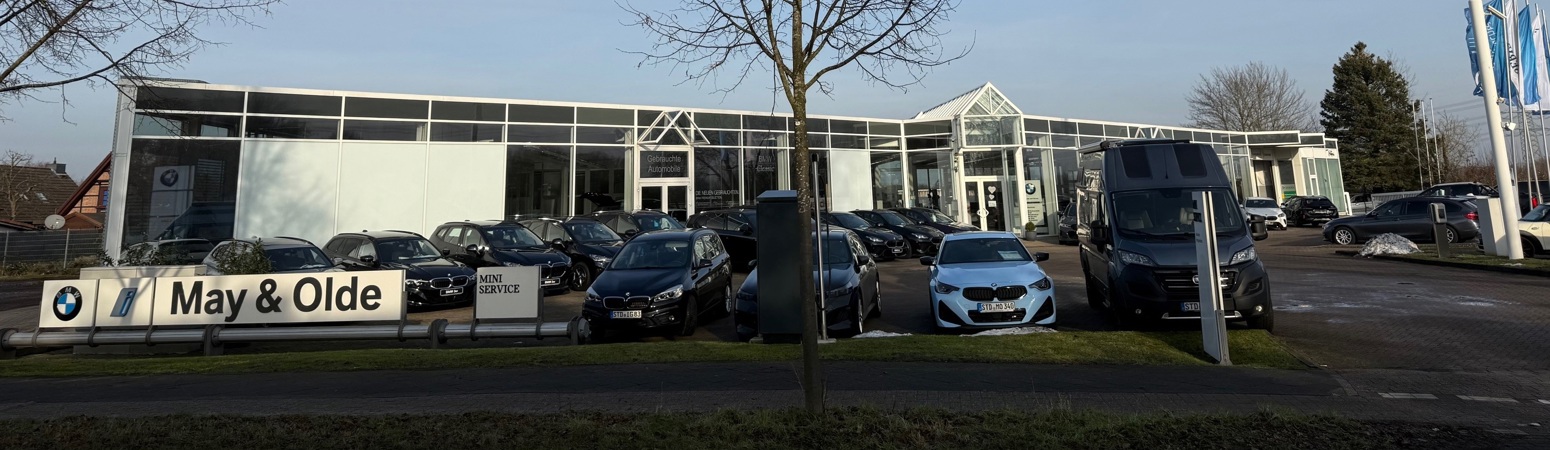 Ein Autohaus mit mehreren BMW-Fahrzeugen, die vor einem modernen Glasgebäude geparkt sind, auf dem ein May & Olde-Schild zu sehen ist.